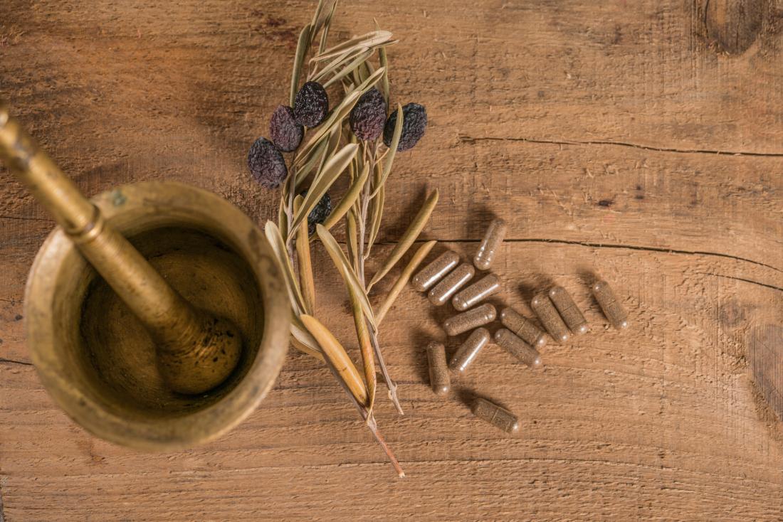 Olive leaf extract supplements on wooden table next to dried olive branch and pestle and mortar.