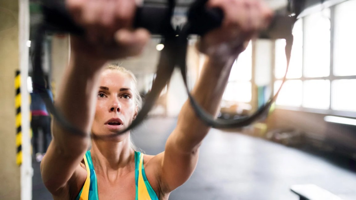 person wearing a yellow and teal tank top reach their arms forward to grasp two TRX straps