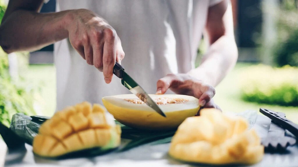 A person slicing a cantaloupe in a kitchen. There are several diced mangos on the kitchen counter.