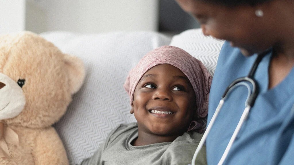 A child wearing a pink head scarf who may have childhood leukemia smiling at someone.