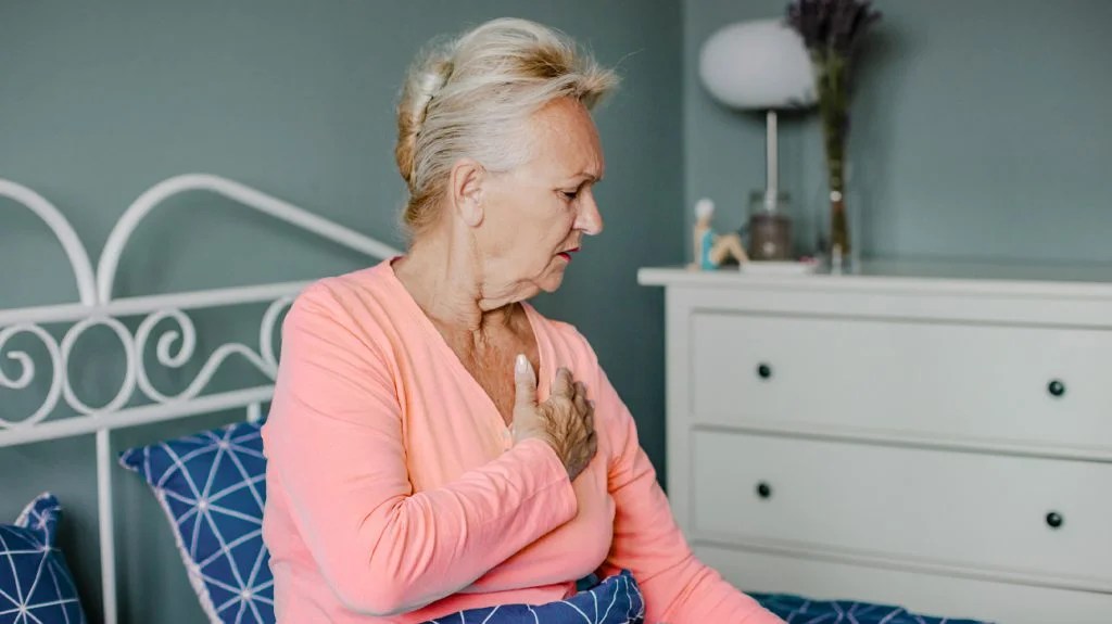 an older woman wearing a pink shirt is sitting on the bed with her hand on her chest