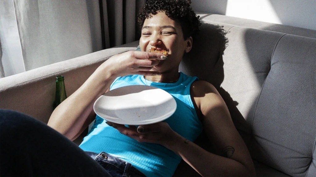 A young woman with diabetes eating food on a gray couch.