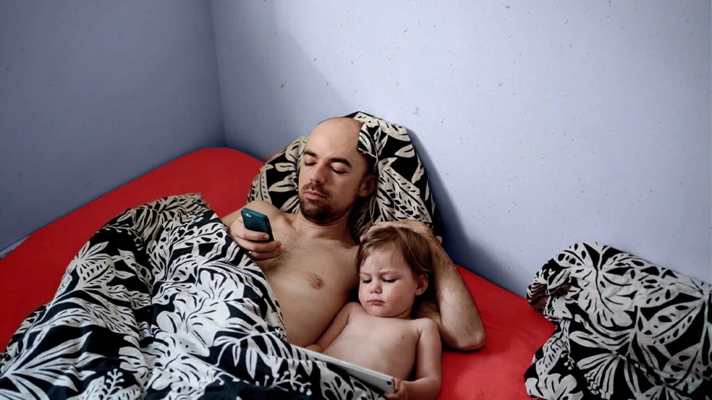 A father and son lying in bed while the father looks at a phone.