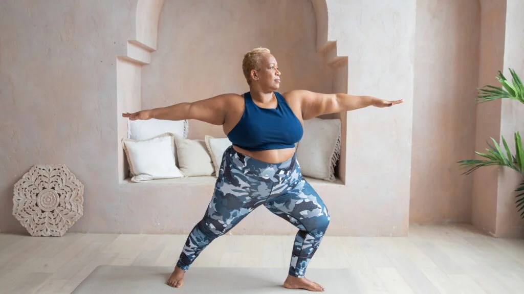 A plus sized woman doing Warrior pose on a yoga mat indoors.