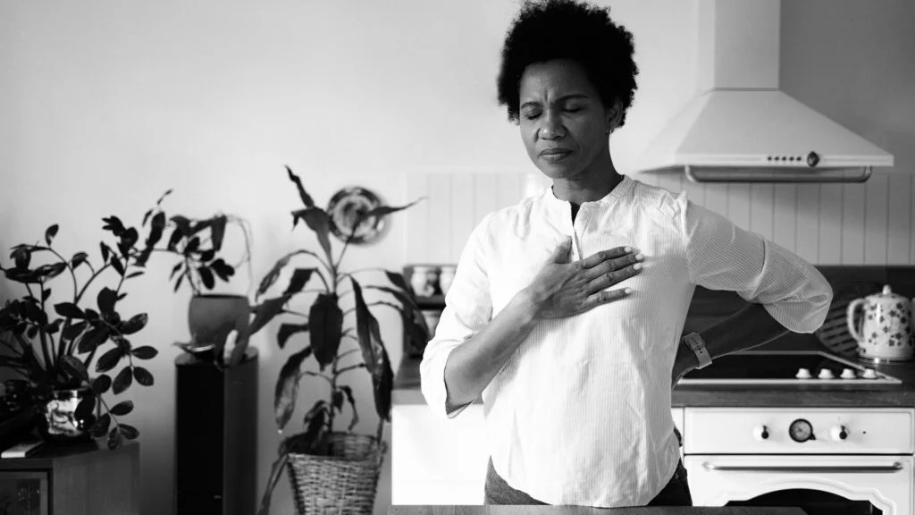 a woman standing in a kitchen with her hand on her chest experiencing pain after breathing deeply -1