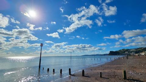 Blue sky with white clouds and the sun to the left with blue sea and beach below