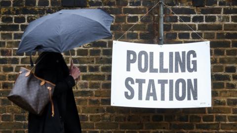 A figure under an umbrella walks past a polling station sign