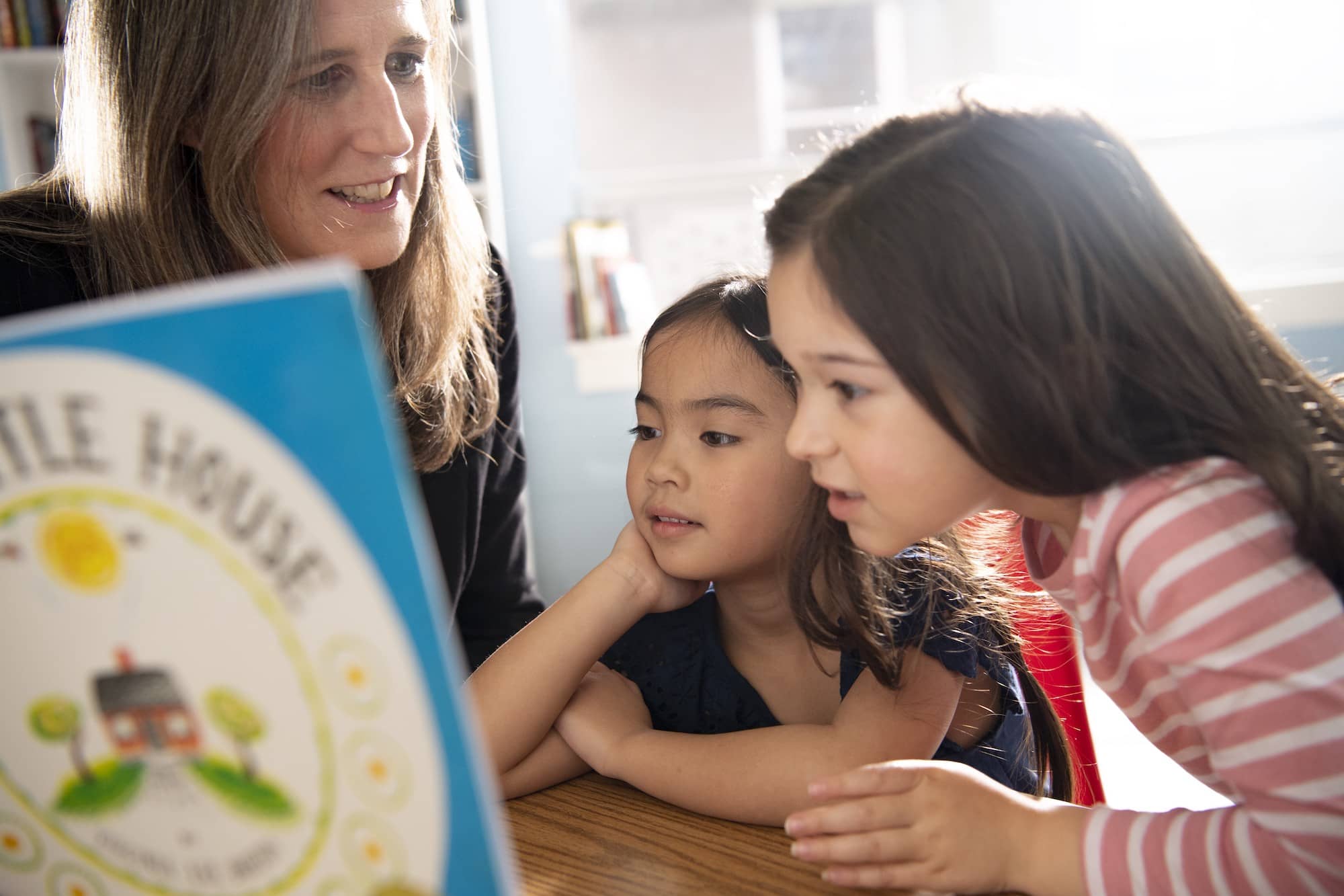 Two girls examine and discuss The Little House by Virginia Lee Barton with their teacher.