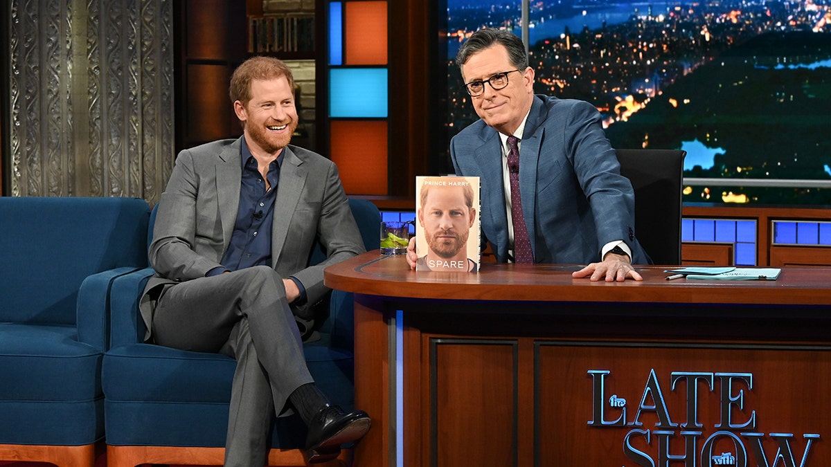 Prince Harry wearing a grey suit and blue shirt smiling sitting next to Stephen Colbert and his memoir Spare