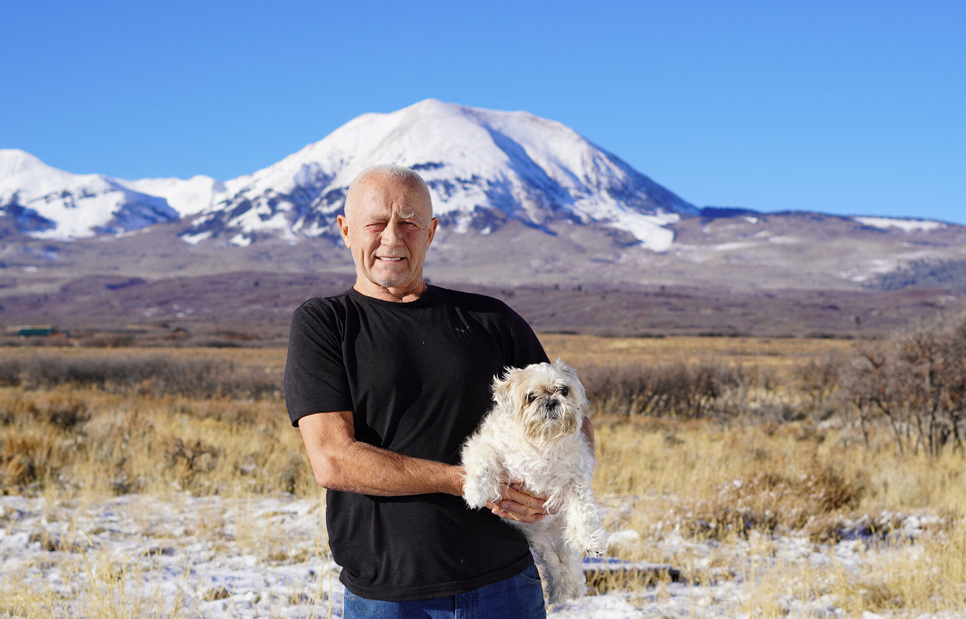 An image of a man smiling while holding a small white dog. There is a landscape behind him with a large, snow-capped mountain in the distance.