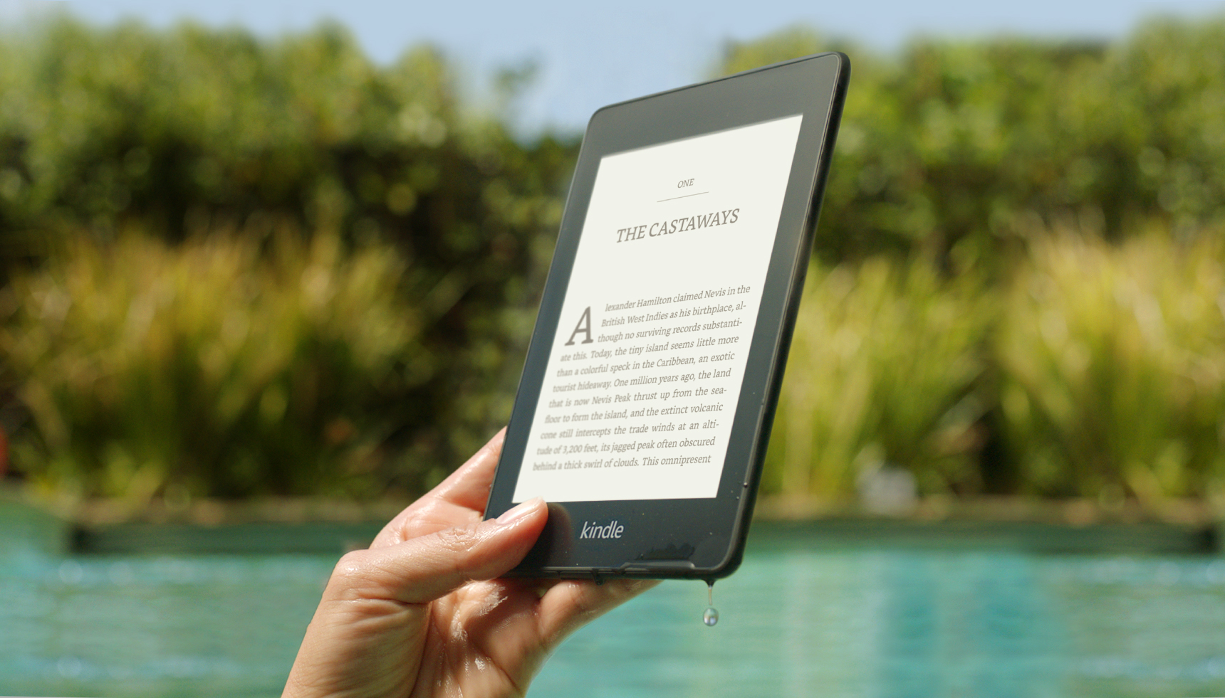 Someone holding their Amazon Kindle Paperwhite by a swimming pool. There is water dripping from the corner of the device. 