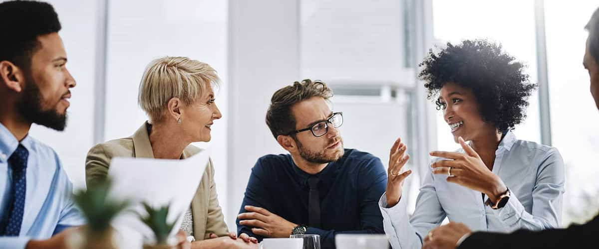 A group of business people sits around a table and listens while a woman speaks.