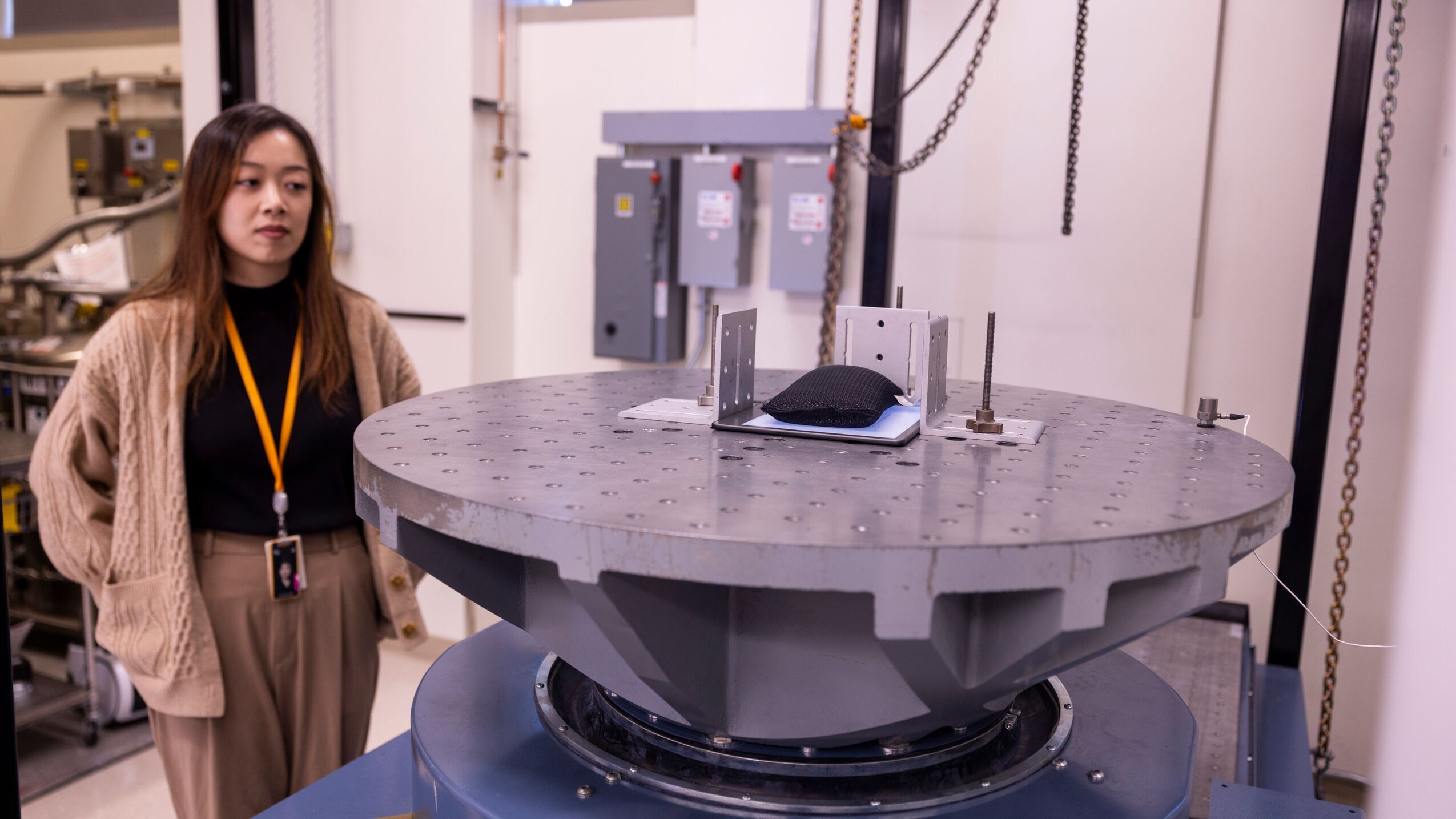 An image of a woman standing in front of a large metal plate with a Kindle device on top of it. She is looking at the machine which is built to test Kindle devices. 