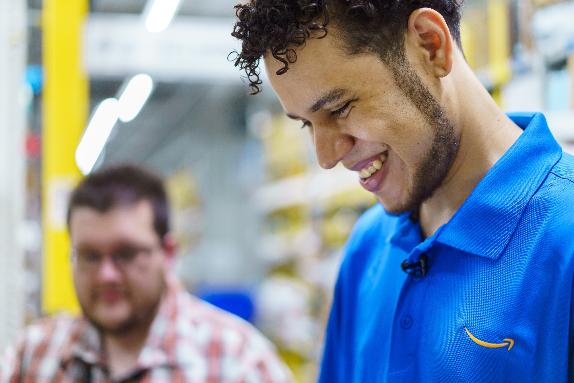 A smiling man is photographed from the side. He wears a blue polo shirt with the Amazon smile logo.