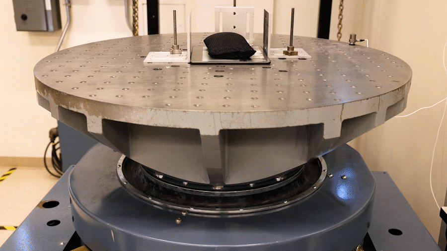 an animated GIF showing a large metal plate with a Kindle device sitting on top of it. There is a weighted bag on top of the device and the plate is vibrating.