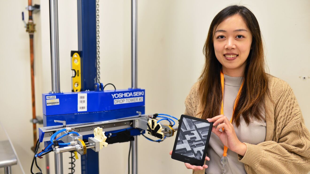 An image of a woman smiling in front of a drop test machine at the Amazon Lab126 Reliability Testing lab in California. She is holding a Kindle device. 