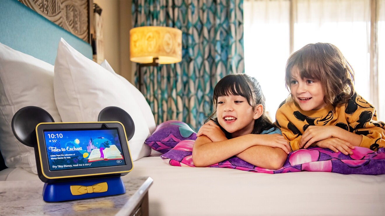 A picture of two kids laying on a hotel bed listening to an Echo smart speaker with Hey Disney! feature. 