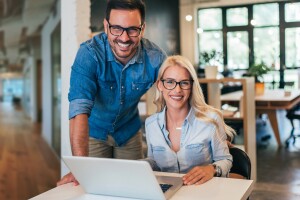 A photo of two business owners. One is sitting at a desk, that has a laptop device on it. The other is standing behind the person who is sitting down.