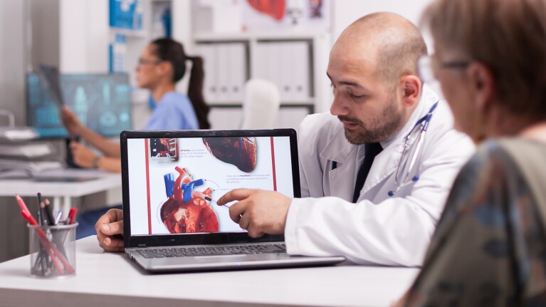 A doctor shows a patient an image describing the functions of the heart on his laptop in a clinic room.