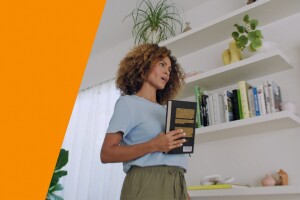 An image of a woman holding a book while talking to Alexa. There is an orange stripe on the left side. 