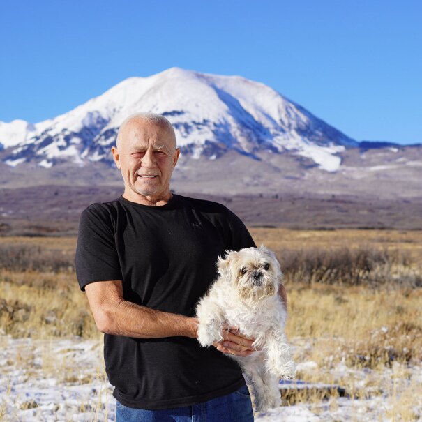 An image of a man smiling while holding a small white dog. There is a landscape behind him with a large, snow-capped mountain in the distance.