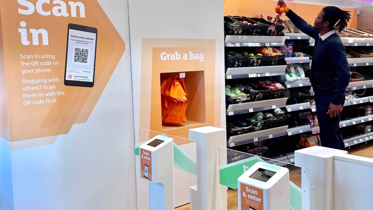 Image of the inside of an Amazon Go store, specifically the entrance. A shopper picks out some fruit from one of the shelves.