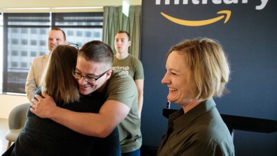 A man wearing a "warriors @ Amazon" shirt hugs a woman. Next to him, another woman smiles. Behind him, two men stand near an Amazon military sign.