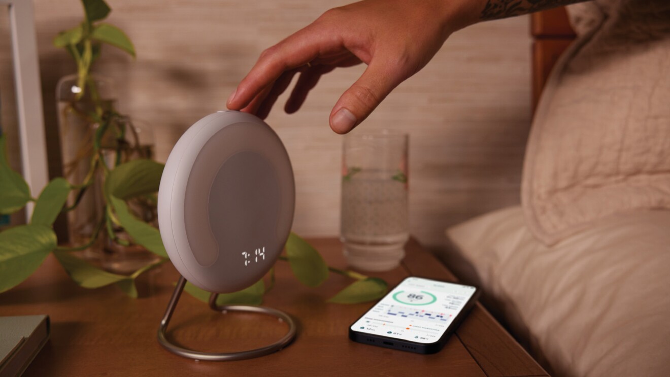 An image of Amazon's Halo Rise sitting on a wooden nightstand with a hand pressing a button from the bed. There is a phone, a glass of water, and a plant also sitting on the nightstand. 