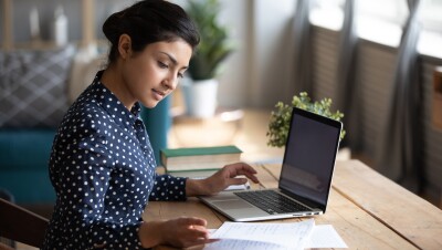 A woman wearing a polka-dotted blouse studies at her desk with her laptop and notes in front of her.