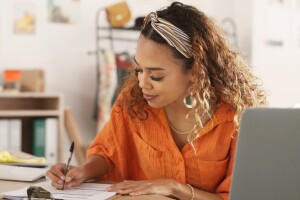 A photo of a business owner writing on a clipboard at a desk. There is also a laptop computer and delivery box next to them.