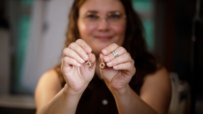 A woman smiles as she holds a pair of earrings towards the camera.