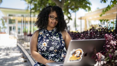 A woman works on a computer in an outdoor setting. She smiles at the camera.