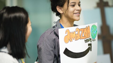 A girl holds up a sign that reads, "Amazon treding global" while another girl smiles and looks at her.