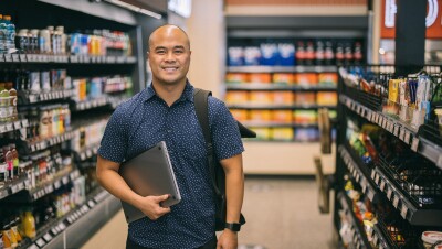 A man holding a laptop computer in one arm smiles at the camera from within an Amazon Fresh grocery store.