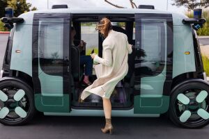 An image showing the Zoox robotaxi opening it's doors on both sides of the vehicle to welcome passengers. There is a passenger entering the vehicle while holding her cellphone and a canned drink. The robotaxi is teal and black and the doors are on a sliding function.