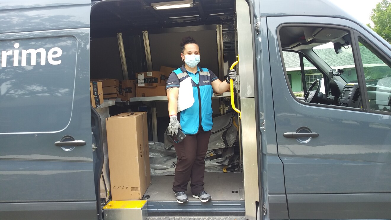 A woman delivery driver wearing a mask stands inside an Amazon van, preparing to deliver customer packages. 