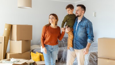 A photo of a family of three standing in a living room of a home. There are moving boxes visible behind them.