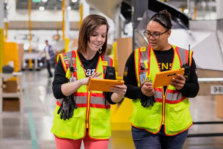 Two Amazon Employees sharing information from their tablets at work. 