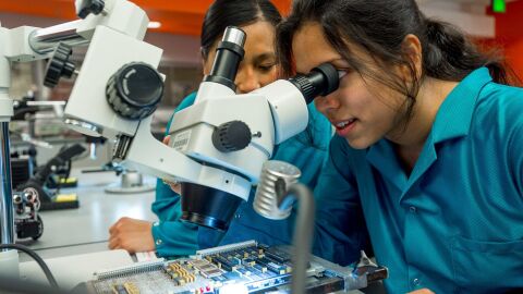 Two women in lab coats using a microscope to examine a circuit board in a laboratory.