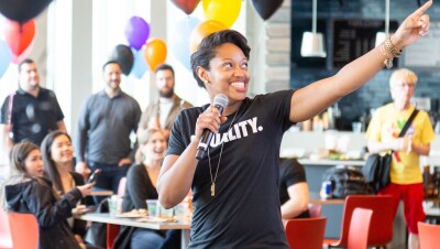 A Black woman wearing a shirt that says "Equality" speaks into a microphone while gesturing off camera. She's speaking to a brightly decorated room full of people.
