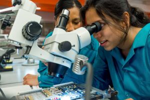 Two women in lab coats using a microscope to examine a circuit board in a laboratory.