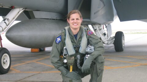 An image of Sarah Rhoads in her pilot gear kneeling in front of a large plane and smiling for a photo.