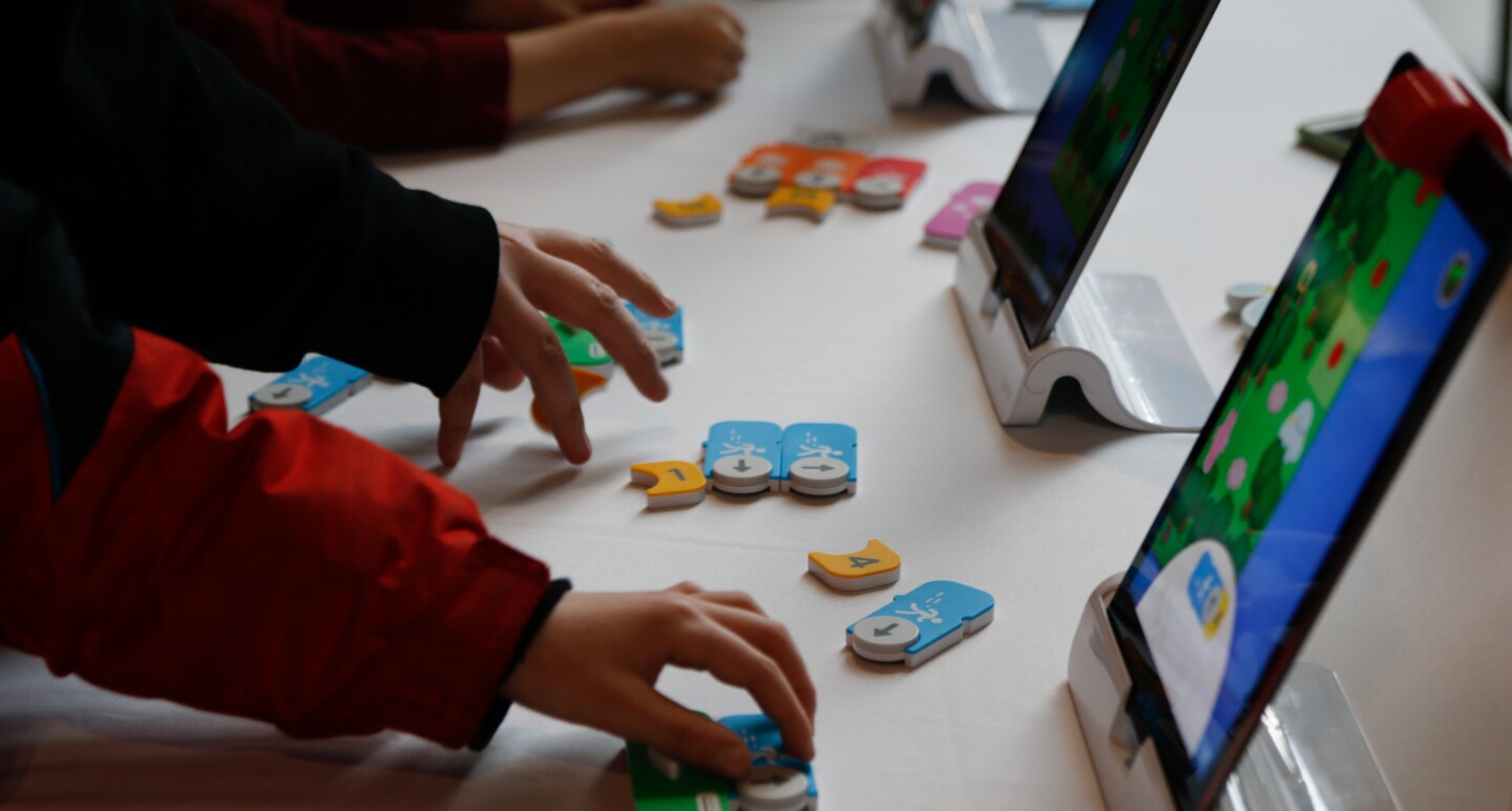 Children hold wooden game pieces, with a tablet placed on the table in front of them. 