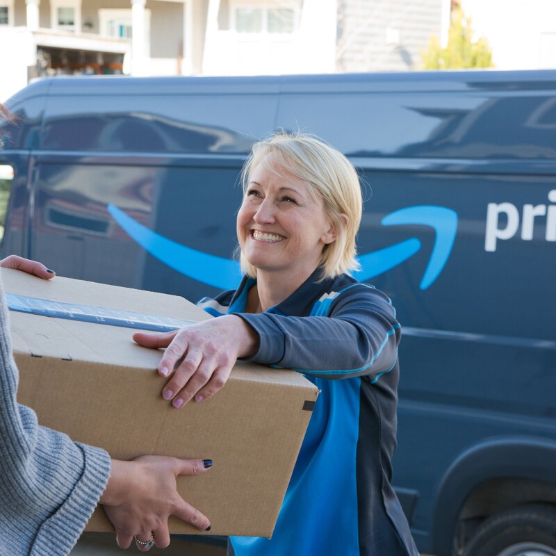 A woman delivers a box to another woman. The delivery driver stands in front of an Amazon DSP van. 