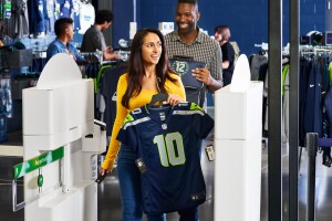 A photo of a customer holding a new Seahawks jersey, walking out of a Seahawks team store, that uses Just Walk Out technology.