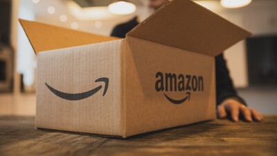 A smiling woman, slightly out of focus, stands behind an open shipping box emblazoned with the Amazon logo, which is resting on her kitchen table.