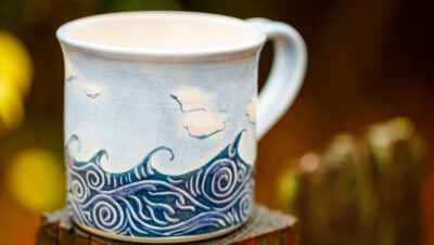 Ceramic mug with painted waves and clouds sits on a wood display pillar.