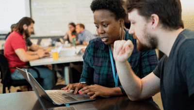 A white male employee follows along as a Black female employee walks him through work on her laptop.