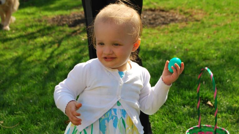 A 22 month old baby girl walking outside holding an easter egg with a basket nearby.
