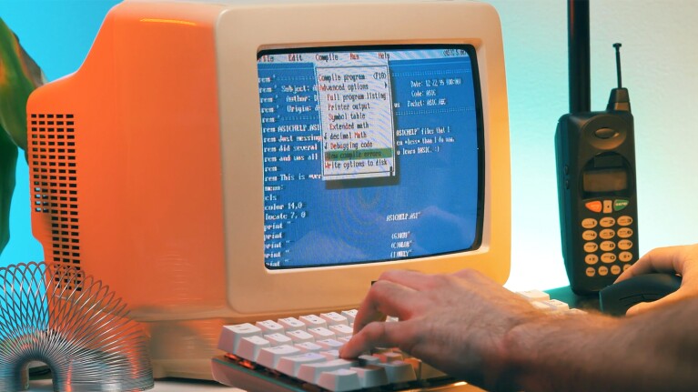 Image of an 80s retro computer with two hands typing on the keyboard. The computer screen displays code and an old phone and slinky are on either side.